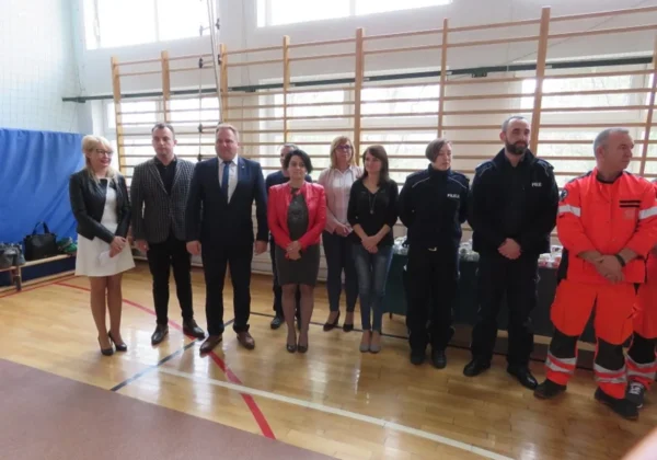 Grupa osób w różnym stroju (urzednicy, policjanci i pracownicy służb ratowniczych) stoi w sali gimnastycznej podczas kampanii społecznej „Jesteś widoczny, jesteś bezpieczny” w Bulkowie.