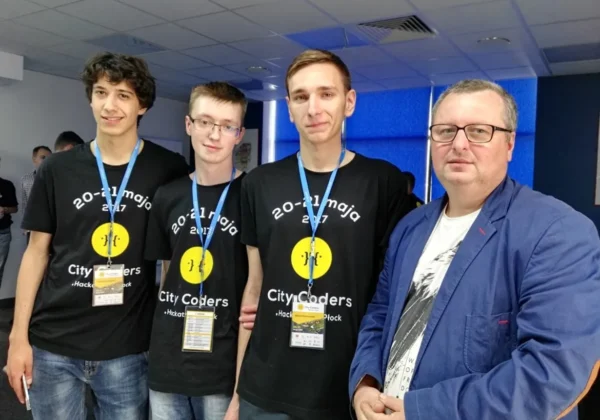 Czterech uczestników Hackathon City Coders stoi razem w sali konferencyjnej: trzech młodych mężczyzn w czarnych koszulkach z żółtym logiem i identyfikatorami oraz dorosły mężczyzna w niebieskiej marynarce.