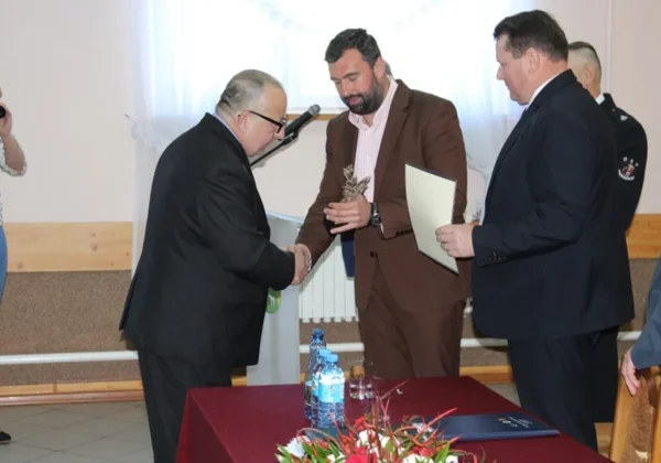 Dwóch mężczyzn w garniturach ściska sobie ręce podczas uroczystości w sali; jeden z nich trzyma statuetkę, obok trzymany jest dokument, a w tle stoją inni uczestnicy przy stole z kwiatami i napojami.