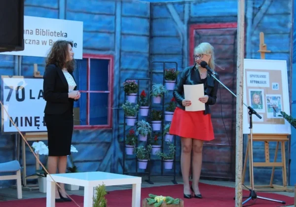 Na scenie dwie kobiety podczas jubileuszu 70-lecia Biblioteki Publicznej w Bulkowie: jedna przemawia do mikrofonu, druga stoi obok, w tle dekoracje, kwiaty i plansza z informacją o spotkaniu autorskim.