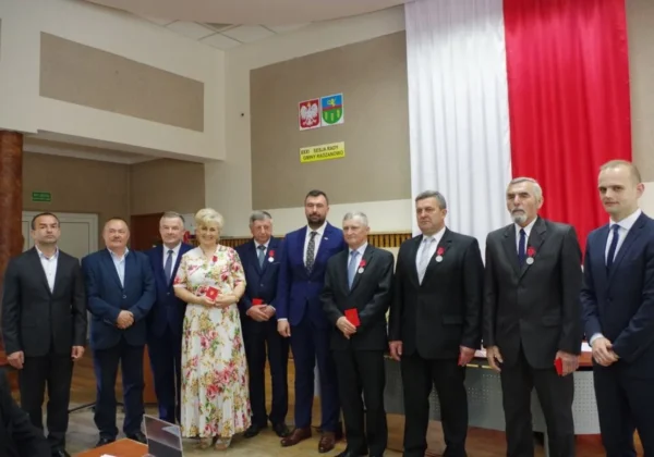 Grupa osób w garniturach i kobieta w kwiecistej sukni stoi na scenie podczas uroczystości wręczania odznaczeń Zasłużonych dla Powiatu Płockiego w Radzanowie, z flagą państwową w tle.
