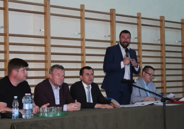 Grupa mężczyzn siedzi za stołem podczas konferencji samorządowej, a jeden z nich stoi i przemawia do mikrofonu.