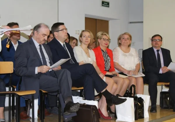 Grupa dorosłych w garniturach i elegancyjnych ubraniach siedzi w rzędzie na sali konferencyjnej, trzymając dokumenty i torby podczas formalnego wydarzenia.