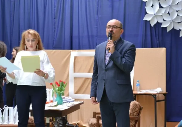 Na scenie przy niebieskiej kurtynie mężczyzna w garniturze przemawia do mikrofonu, podczas gdy kobieta trzyma kartki i czyta je, otoczona prezentami i kwiatami.