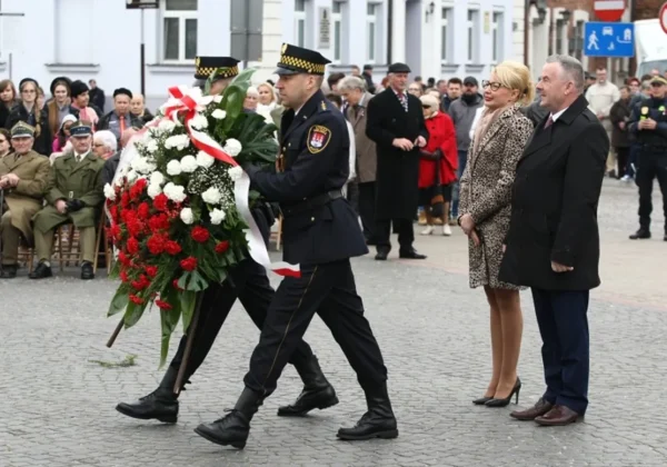 Uroczystość z okazji 226. rocznicy Konstytucji 3 Maja: funkcjonariusz w mundurze niesie biało‑czerwony wieniec przed zgromadzonymi mieszkańcami i urzędnikami, w tle stoją inni uczestnicy.