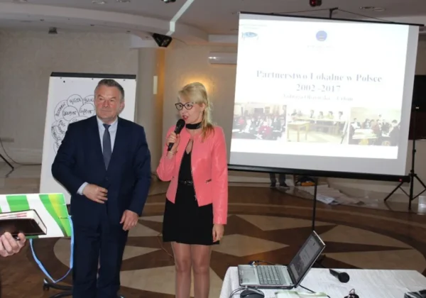 Dwóch prelegentów w sali konferencyjnej prezentuje start Partnerstwa Lokalnego w powiecie płockim; mężczyzna w garniturze i kobieta z mikrofonem stoją obok ekranu z prezentacją.