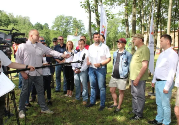 Grupa osób w parku podczas konferencji prasowej z mikrofonami i kamerą, w otoczeniu drzew, promuje spływ kajakowy na Skrwie i Ziemi Płockiej.