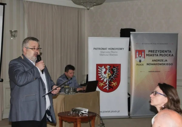 Mężczyzna w garniturze przemawia do mikrofonu podczas Dni Integracji i Aktywności Osób Niepełnosprawnych, na tle bannerów z herbem powiatu płockiego i informacjami o prezydencie Miasta Płocka.