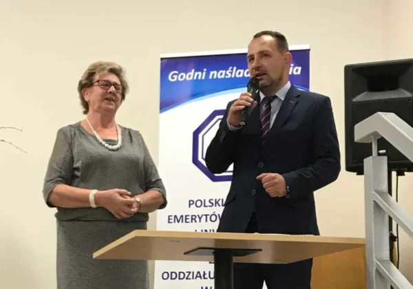 Dwóch prelegentów na scenie podczas uroczystości z okazji Światowego Dnia Inwalidy: mężczyzna w garniturze przemawia do mikrofonu, obok niego stoi kobieta w szarej sukience z perłową biżuterią, w tle baner i głośnik.
