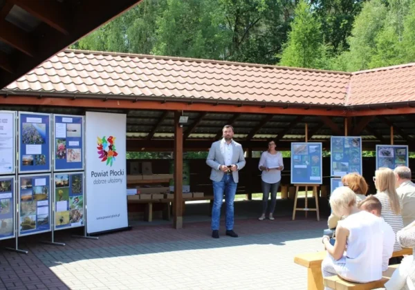 Na zdjęciu dwie osoby stoją na plenerowej scenie pod drewnianym zadaszeniem podczas wręczania nagród w konkursie fotograficznym, w tle widoczna jest wystawa fotografii na tablicach i banner z logo Powiatu Płockiego, a publiczność siedzi na ławkach.