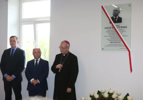 Na odrestaurowanej siedzibie delegatury MUW w Płocku trzy osoby w formalnych garniturach (dwóch mężczyzn i duchowny) stoją przy tablicy pamiątkowej z wizerunkiem nieokreślonej osoby i czerwono-białą wstęgą, przed nimi leży bukiet białych róż.