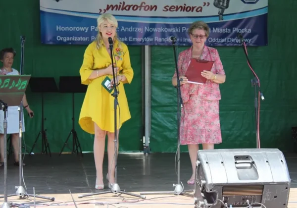 Na scenie podczas III Festiwalu Amatorskich Zespołów „Mikrofon Seniora” dwie kobiety mówią do mikrofonów; jedna w żółtej sukience, druga w różowej, kwiecistej sukience, z bannerem w tle.