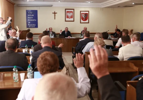 Uczestnicy XXV sesji Rady Powiatu w Płocku podnoszą ręce podczas głosowania w sali obrad, a w tle widoczne są krzyż i dwa herby na ścianie.