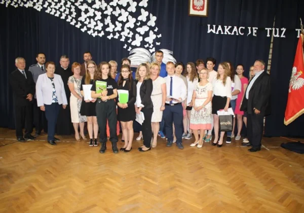 Grupa uczniów i nauczycieli na scenie podczas zakończenia roku szkolnego w ZS w Wyszogrodzie, z dekoracją białych motyli i napisem „WAKACJE TUŻ TUŻ” w tle.
