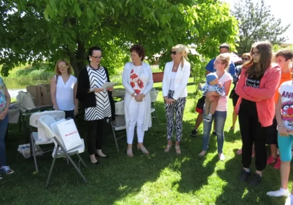 Grupa dorosłych i dzieci stoi na zielonej trawie pod drzewem podczas pikniku z okazji Dnia Rodzicielstwa Zastępczego i Dnia Dziecka.