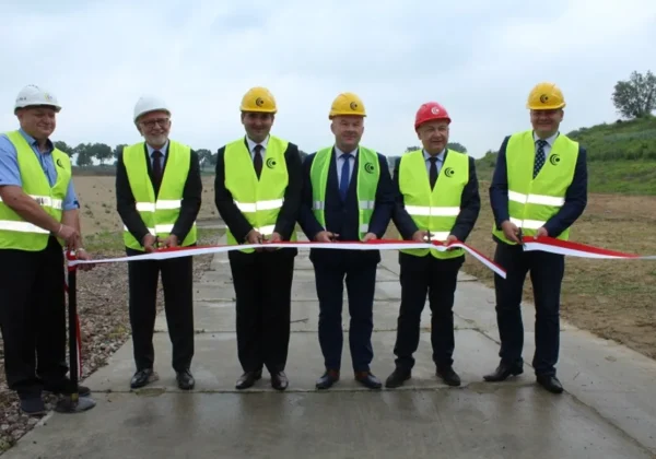 Grupa sześciu mężczyzn w kaskach i kamizelkach odblaskowych przecina czerwono-białą wstęgę podczas inauguracji inwestycji w Kobiernikach.