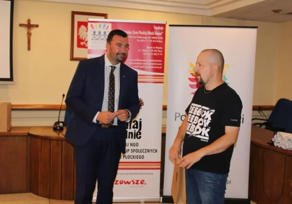 Dwóch mężczyzn w sali konferencyjnej podczas ceremonii, jeden w garniturze wręcza nagrodę drugiemu, a w tle stoją banery.