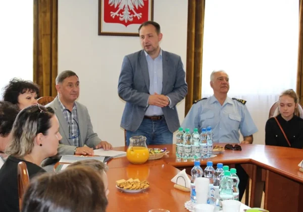 Mężczyzna stojący przy stole konferencyjnym przemawia podczas wręczania nagród laureatom konkursu plastycznego, w obecności innych uczestników i z widocznym herbem Polski na ścianie.
