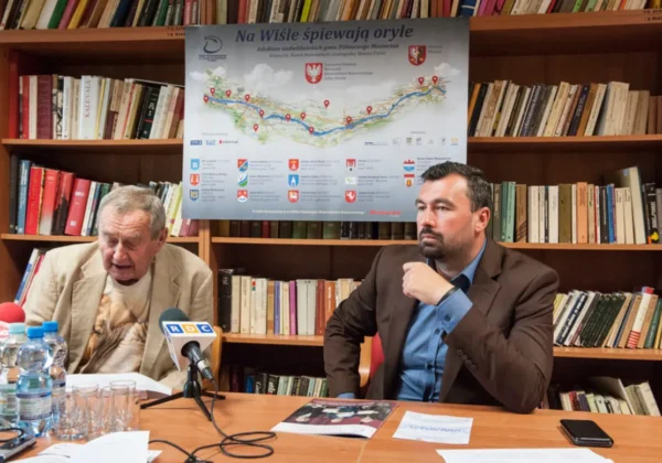 Dwóch mężczyzn podczas konferencji prasowej w bibliotece, z mikrofonami na stole, a w tle plakat promujący Dni Wisły w Wyszogrodzie i wyjątkowy spektakl plenerowy.