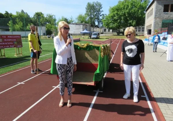 Dwie kobiety stoją na bieżni stadionu podczas VII Powiatowej Olimpiady Integracyjnej; jedna trzyma mikrofon przy dekorowanym wozie, a w tle widać uczestników w białych strojach i baner informacyjny o imprezie.