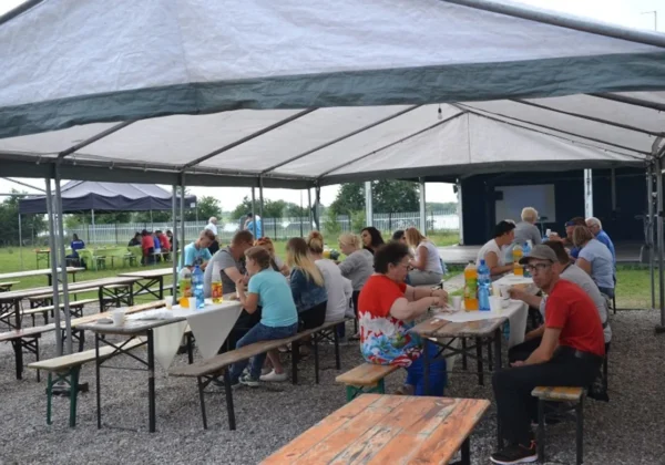 Grupa ludzi siedzi przy długich stołach pod namiotem podczas pikniku integracyjnego, jedzą i piją na świeżym powietrzu, z widoczną sceną w tle.