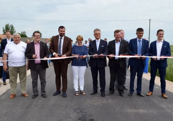 Grupa osób w garniturach i eleganckich strojach stoi na nowo wybudowanej drodze gminnej nr 290207W podczas ceremonii otwarcia, przecinając czerwono-białą wstęgę.