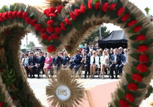 Uczestnicy Dożynek Powiatu Płockiego w Drobinie siedzą podczas ceremonii, a na pierwszym planie widoczny jest kwietno-słomiany wieniec.