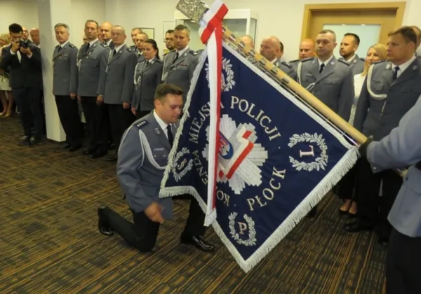 Uroczyste powitanie nowego Komendanta Miejskiego Policji: policjanci w mundurach stoją w sali, a funkcjonariusz klęka i trzyma niebieski sztandar z herbem Policji i napisem Płock.