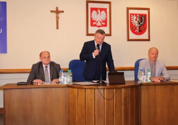 Trzech mężczyzn siedzi przy długim drewnianym stole w sali sesyjnej, przy czym jeden stoi i przemawia do mikrofonu, a w tle wiszą krzyż i dwa oprawione herby.