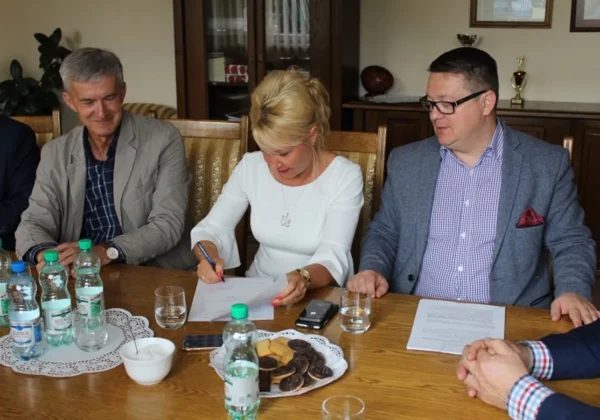 Kobieta podpisuje dokument podczas podpisywania umowy dotyczącej rozbudowy skrzyżowania na drodze powiatowej, w towarzystwie dwóch mężczyzn przy stole z wodą i ciasteczkami.