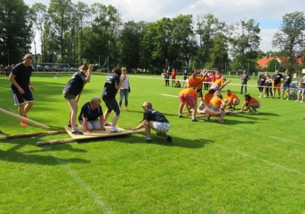 Uczestnicy VII Gminnego Turnieju Sołectw „Sołtysiada 2017” rywalizują na boisku, przenosząc drewnianą platformę po rolkach, podczas gdy widzowie obserwują z boku.
