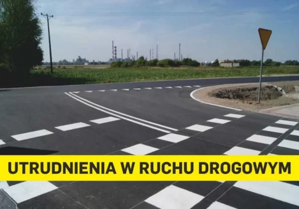Droga z wyraźnym oznakowaniem poziomym i żółtym napisem ostrzegawczym „UTRUDNIENIA W RUCHU DROGOWYM” na tle pól i zabudowań przemysłowych.