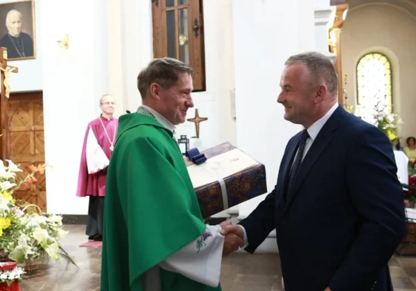 Uroczysta scena w parafii Św. Benedykta w Płocku-Radziwiu: duchowny w zielonym ornacie i mężczyzna w garniturze ściskają dłonie podczas wręczania prezentu we wnętrzu kościoła.