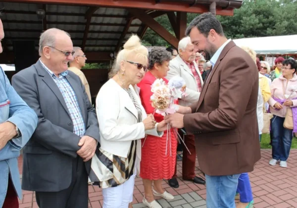 Mężczyzna w brązowej kurtce wręcza bukiet kwiatów starszej kobiecie w białej marynarce podczas wakacyjnego festynu dla seniorów.