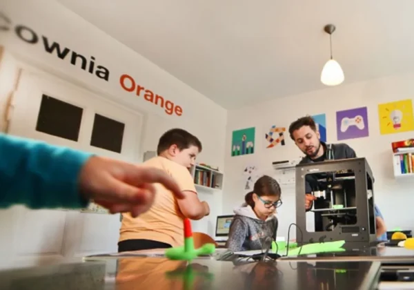Grupa dzieci i dorosły pracuje w nowoczesnej Pracowni Orange, obsługując drukarkę 3D w otoczeniu kolorowych plakatów na ścianach.