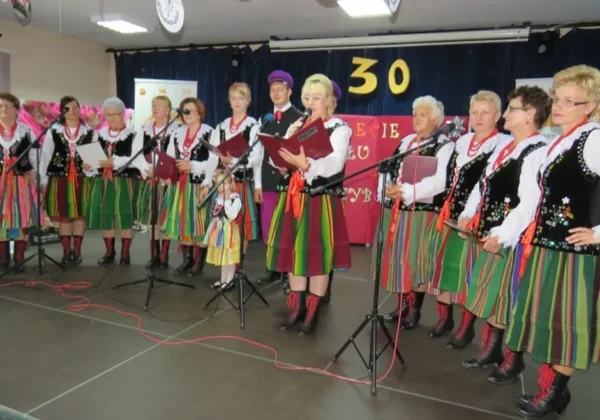 Grupa wykonawców w tradycyjnych, kolorowych strojach ludowych stoi na scenie z mikrofonami i śpiewa podczas jubileuszu 30-lecia Zespołu Ludowego Grzybowianki ze Słubic.