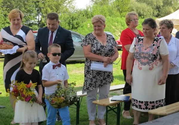 Grupa dorosłych i dzieci na zewnątrz podczas Duninowskiego Święta Plonów i Darów Wisły, z dziećmi w pierwszym planie trzymającymi kwiaty i koszyczek z plonami.