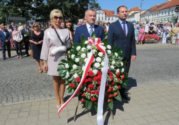 Grupa osób w eleganckich ubraniach stoi z okazałym wieńcem z białych i czerwonych kwiatów ozdobionym wstęgą w barwach narodowych, podczas uroczystości Święta Wojska Polskiego.