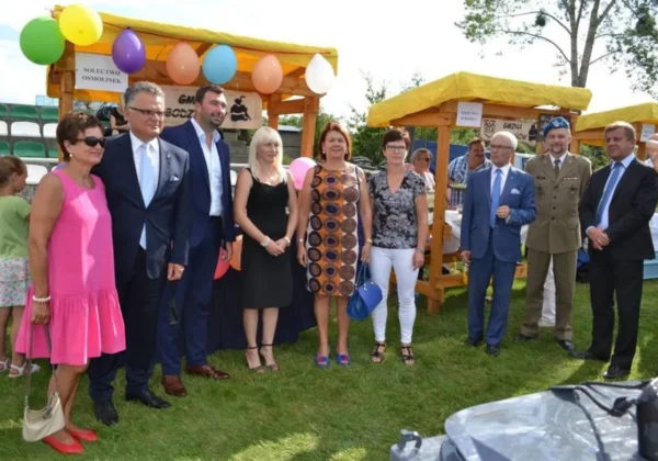 Grupa osób w eleganckich strojach stoi na trawie przed drewnianymi pawilonami ozdobionymi balonami podczas Dni Bodzanowa.
