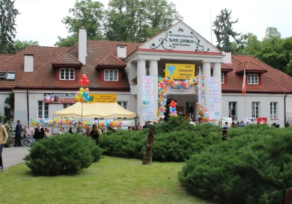 Plenerowy festyn przed budynkiem Domu Pomocy Społecznej w powiecie płockim, z balonami, stoiskami i ludźmi przed wejściem.
