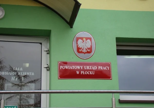 Widok na zewnętrzną część budynku Powiatowego Urzędu Pracy w Płocku, z emblematem Polski i tablicą z nazwą placówki na zielonej ścianie.