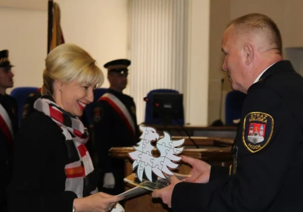 Podczas obchodów 25-lecia Straży Miejskiej w Płocku kobieta odbiera od strażnika miejskiego nagrodę w formie puzzli tworzących drzewo.