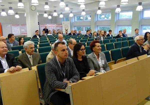 Uczestnicy konferencji „Młodzi dla Techniki 2017” siedzą w sali konferencyjnej, w pierwszym rzędzie przy drewnianych biurkach, uważnie obserwując prezentację.