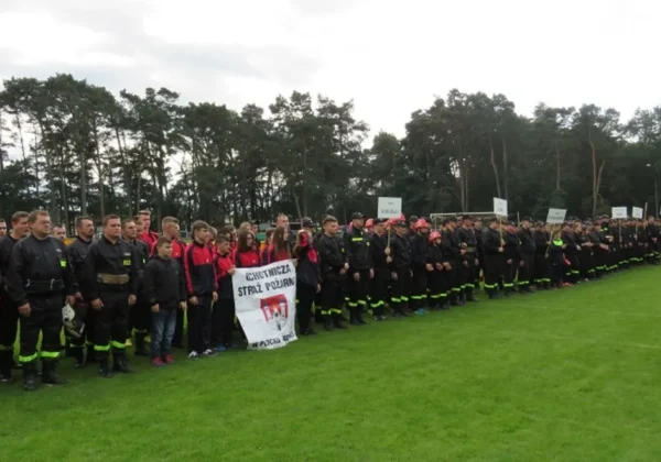 Duża grupa strażaków OSP i młodzieżowych drużyn pożarniczych stoi w rzędzie na zielonej murawie podczas X Powiatowych Zawodów Sportowo-Pożarniczych.