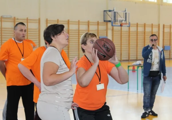 Grupa uczestników sportowo‑muzycznych zmagań w hali w Radzanowie, w pomarańczowych koszulkach, z kobietą trzymającą piłkę do koszykówki na pierwszym planie i prowadzącym z mikrofonem w tle.