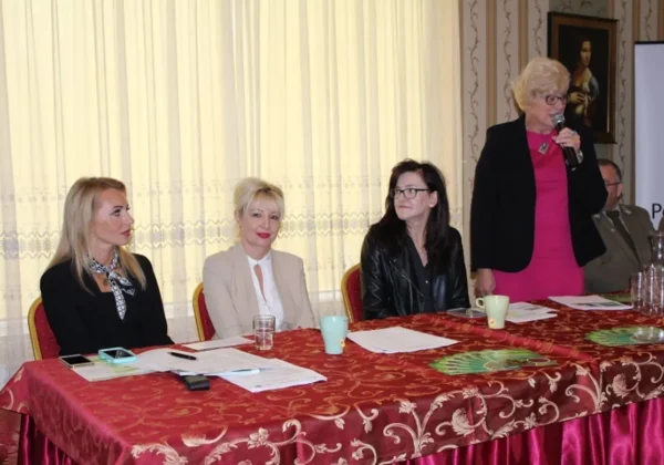 Kobieta stojąca z mikrofonem przemawia podczas Ekokonferencji „Chrońmy naturę” w Wyszogrodzie, podczas gdy trzy inne kobiety siedzą przy długim stole przykrytym czerwonym obrusem.