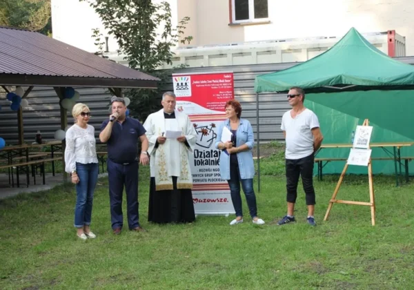 Grupa osób, w tym duchowny w ornacie, stoi na trawie obok namiotu i baneru podczas plenerowego wydarzenia promującego zdrowy, weekendowy tryb życia w Nowym Duninowie.