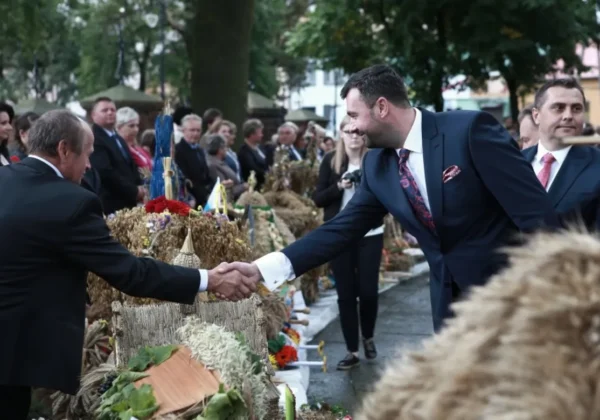 Dwóch mężczyzn w garniturach ściska sobie ręce na tle dekoracji z ziaren i wieńców podczas Dożynek Powiatu Płockiego 2017.