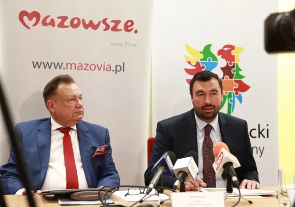 Dwóch mężczyzn w garniturach siedzi przy stole podczas konferencji prasowej o milionach na drogi powiatowe; w tle widoczne banery Mazowsza.