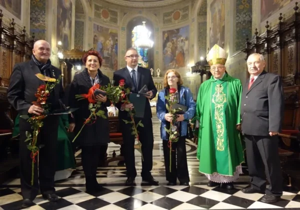 Grupa sześciu osób stoi w bogato zdobionym kościelnym wnętrzu, wśród nich duchowny w zielonej szacie trzyma kwiaty i nagrody.